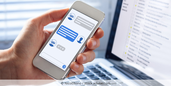 A hand holding a smartphone that shows the conversation with a chatbot and a laptop screen in the background.