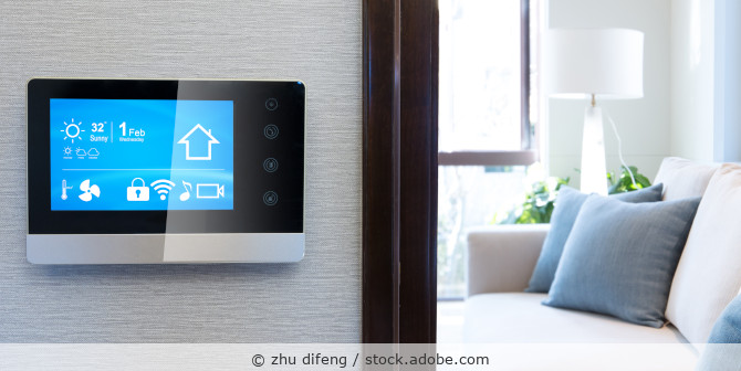 Touchscreen for smart home control mounted on a wall with a living room visible in the background.